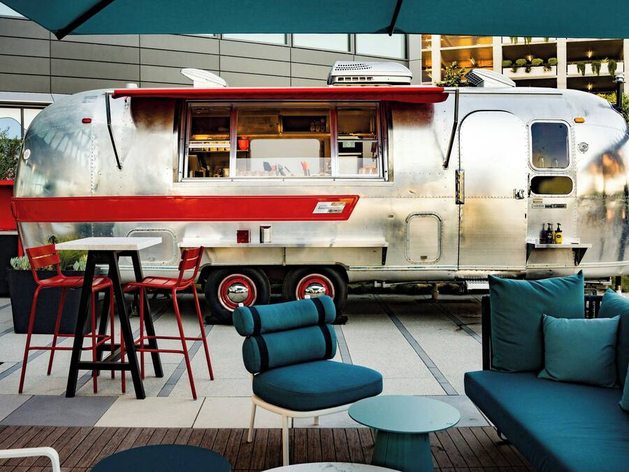 A vintage food truck with a red and white exterior parked in an outdoor seating area with blue furniture and a modern building in the background.
