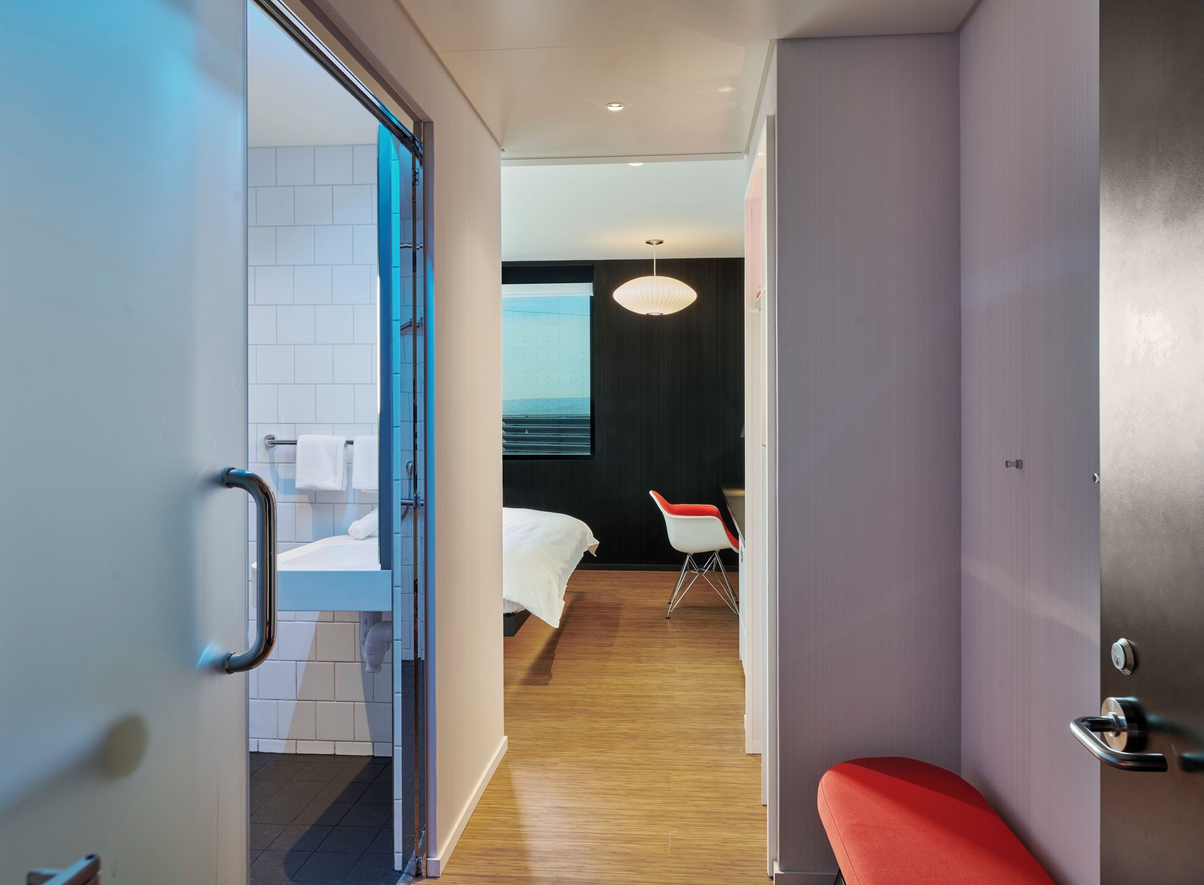 A modern hotel room with a wooden floor, a bed, a red chair, and a view of the sea through a window.