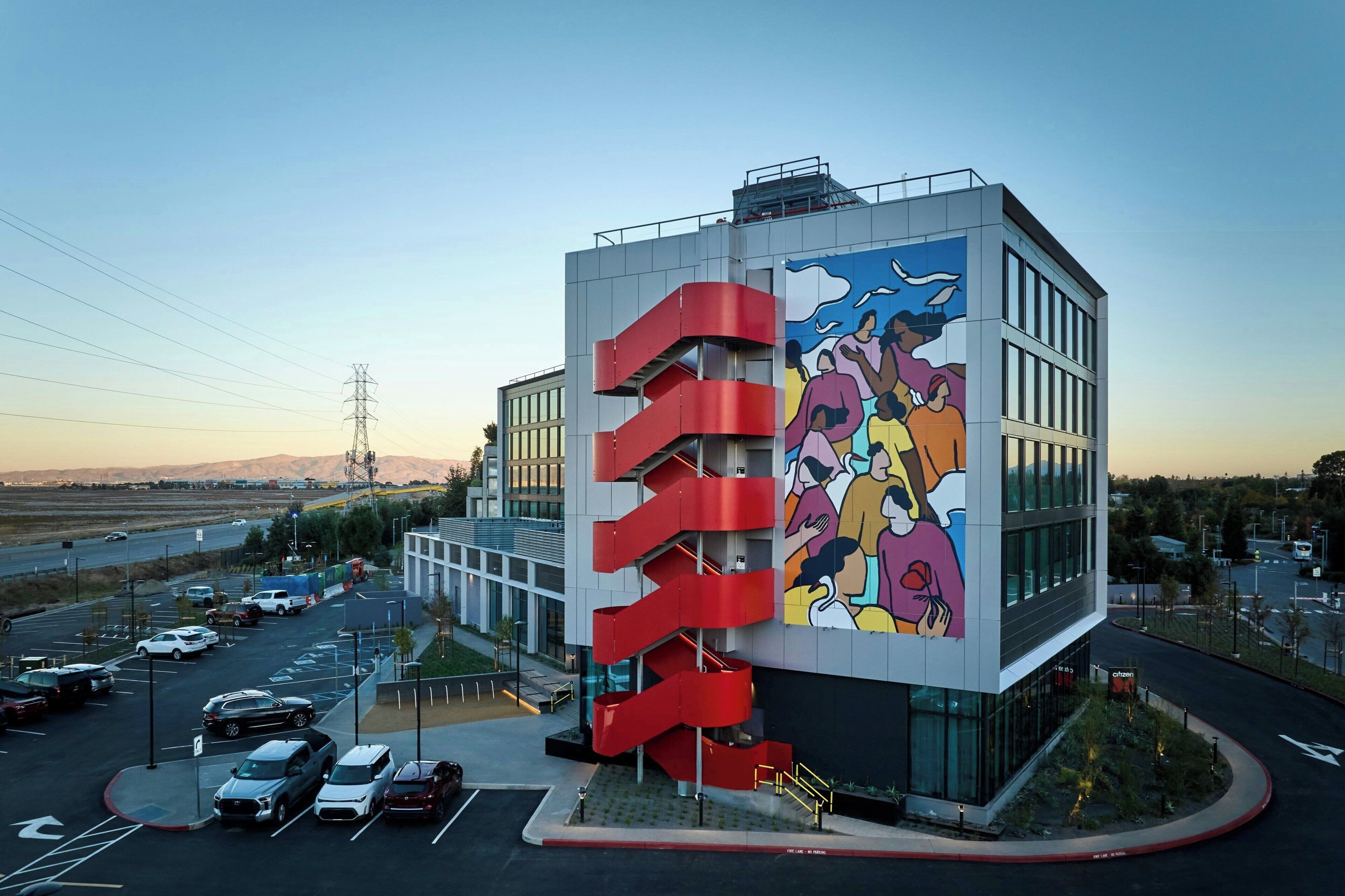 Un edificio moderno con una escalera espiral roja singular y un mural colorido en la fachada.