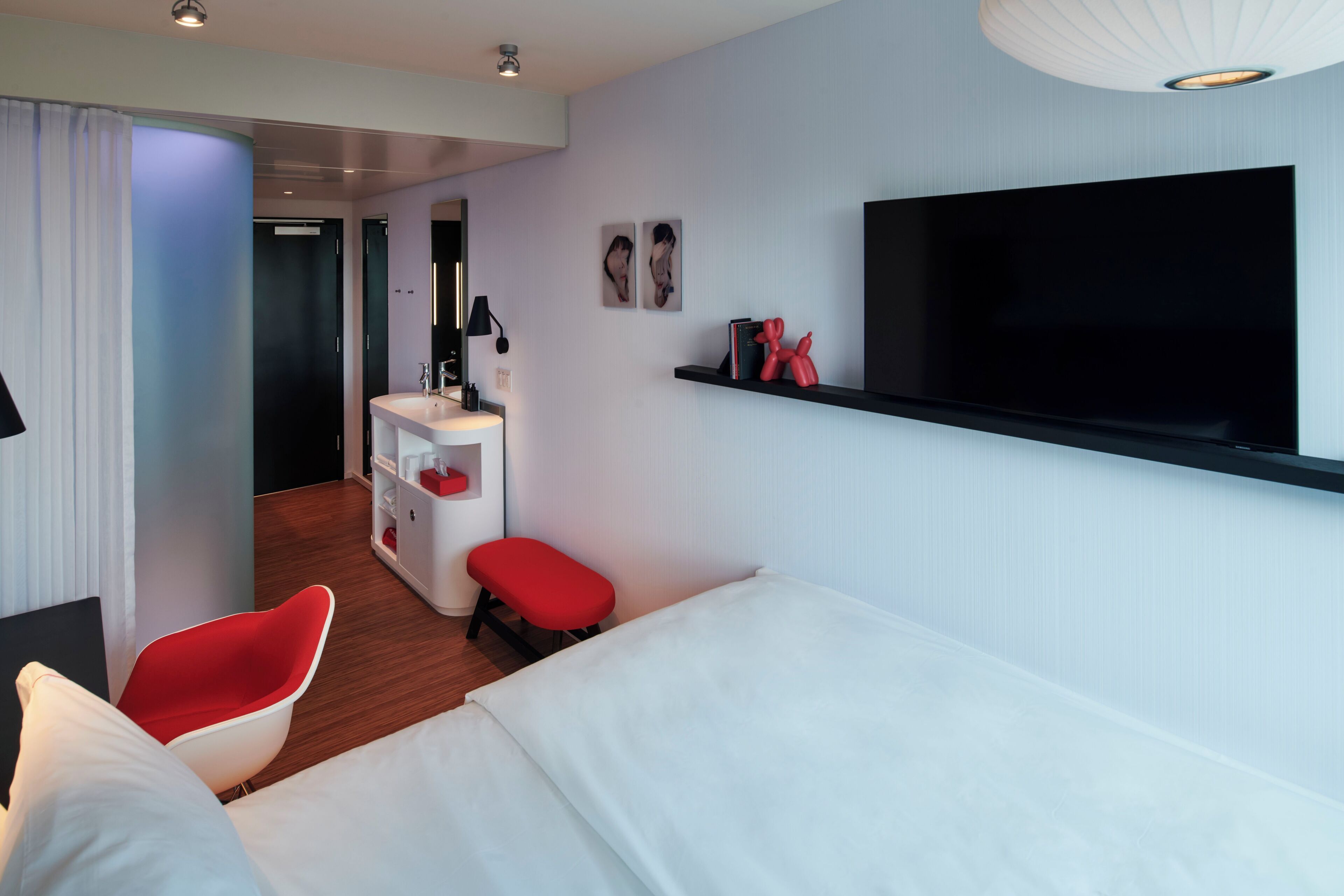 Habitación de hotel moderna con una cama, una silla roja, un escritorio pequeño con un espejo, un televisor montado en la pared y una escultura roja en una estantería.