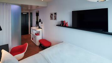 Habitación de hotel moderna con una cama, una silla roja, un escritorio pequeño con un espejo, un televisor montado en la pared y una escultura roja en una estantería.