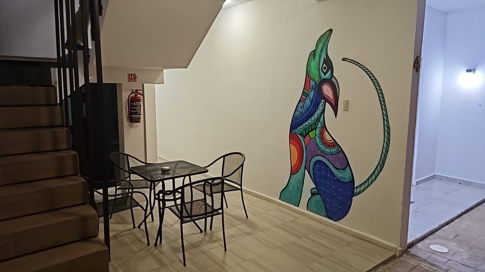 A staircase leading to a room with a mural of a colorful bird, a small table with chairs, and a fire extinguisher on the wall.
