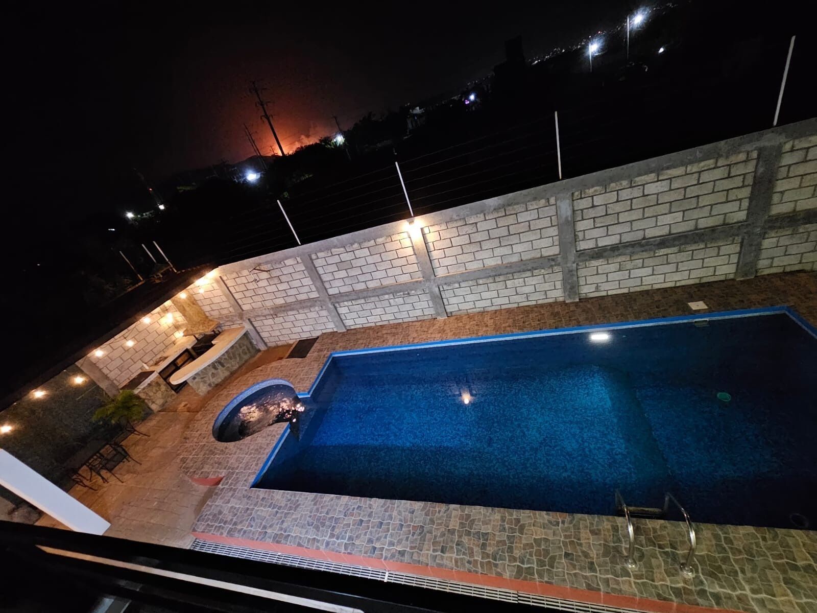 Una piscina con pared de ladrillo y terraza de piedra, rodeada de iluminación exterior.
