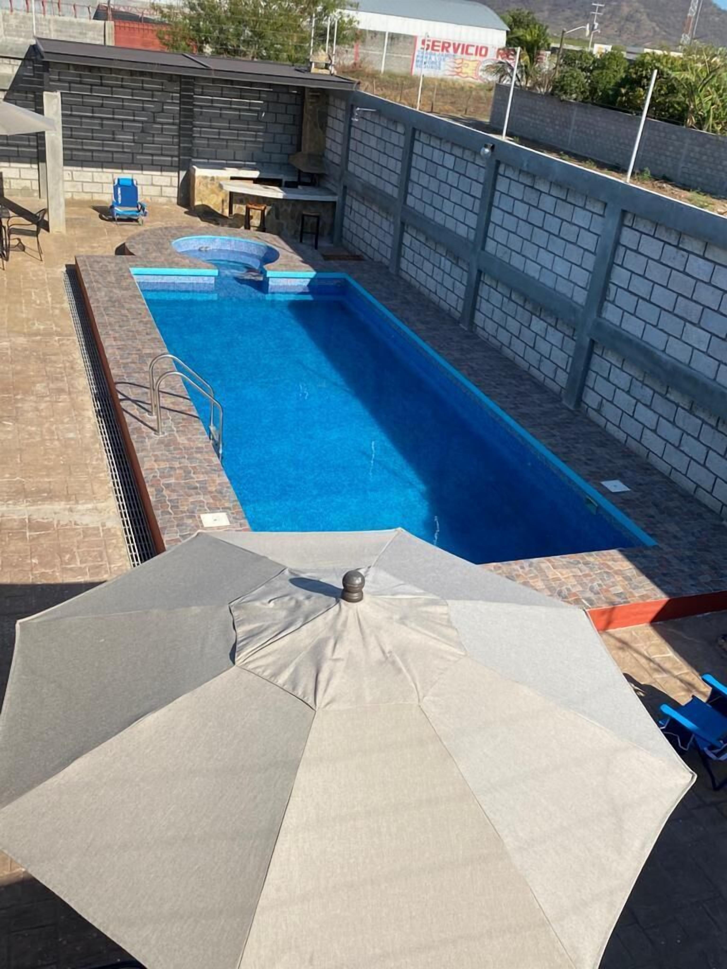 Una piscina rectangular con revestimiento azul, rodeada por un muro de ladrillo y una cerca blanca. Hay una sombrilla blanca y una silla azul cerca de la piscina.