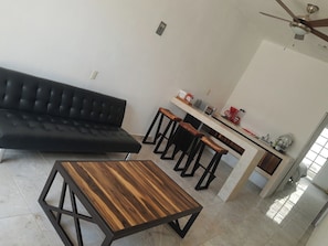 A modern living room with a black sofa, a wooden coffee table, and a kitchen area with a bar stool and a coffee machine. - Hotel Boutique Alebrijes Salina Cruz (Salina Cruz)