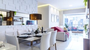 Un comedor moderno con una mesa blanca para cuatro, un sofá con cojines y vistas a la ciudad a través de amplias ventanas.