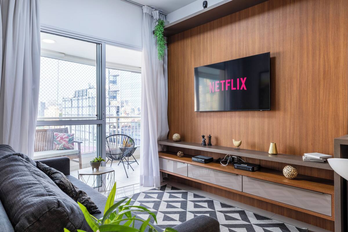 Ein modernes Wohnzimmer mit einem großen Flachbildfernseher an einer Holzwand, einem grauen Sofa, einem kleinen Beistelltisch und einem gemusterten Teppich.