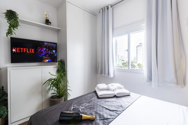 A hotel room with a bed, a TV displaying the Netflix logo, a bottle of champagne, and a window with curtains.