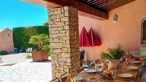 An outdoor dining area with a stone pillar, red umbrellas, and a long wooden table set for a meal.