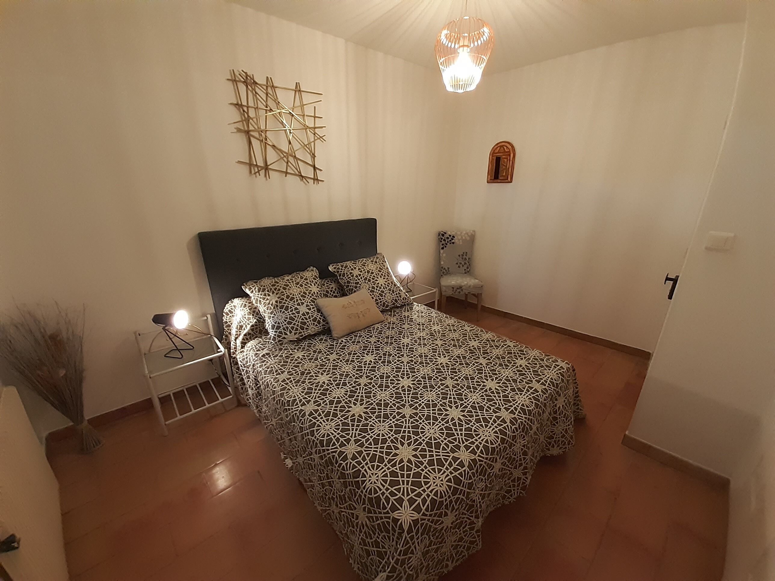 A bedroom with a patterned bedspread, a nightstand with a lamp, a chair, and a decorative wall piece.