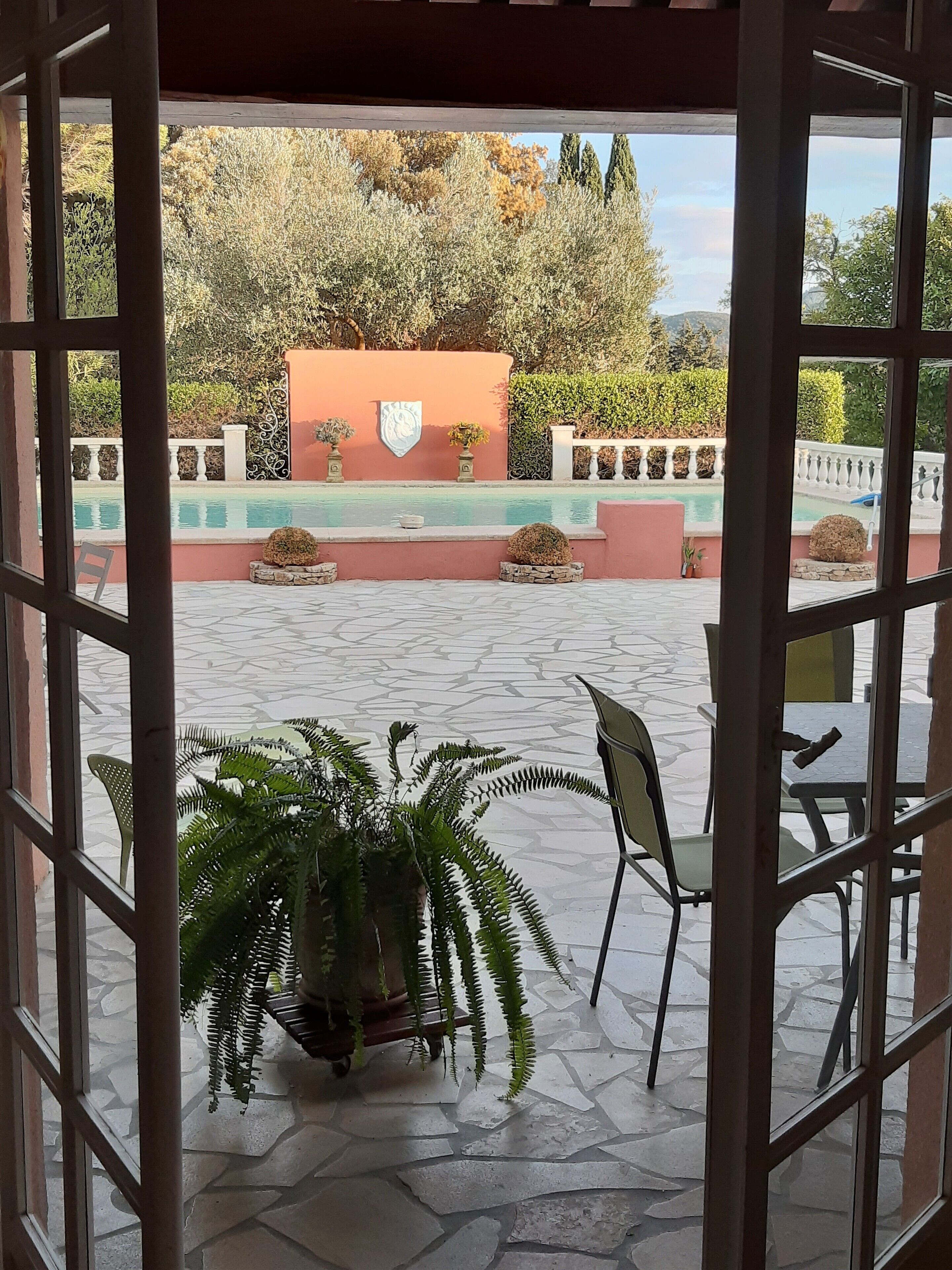 Un patio al aire libre con suelo de piedra, una piscina y vistas a un jardín ajardinado con árboles y un muro.