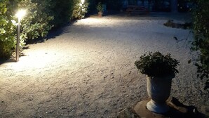 A nighttime garden scene with a gravel path, illuminated by street lamps, surrounded by trees and a potted plant.