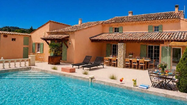 A poolside area with a clear blue pool, outdoor seating, and a building with a tiled roof.