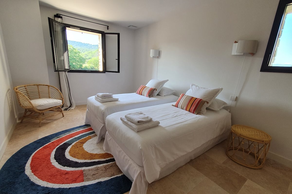 Un dormitorio con dos camas, una silla de ratán, una mesita redonda y un tapete colorido.