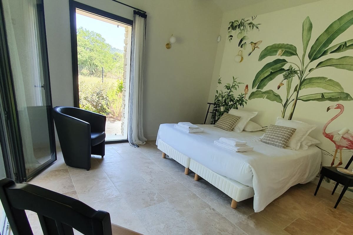 Un dormitorio con cama, una silla, una mesa y un ventanal con cortinas.