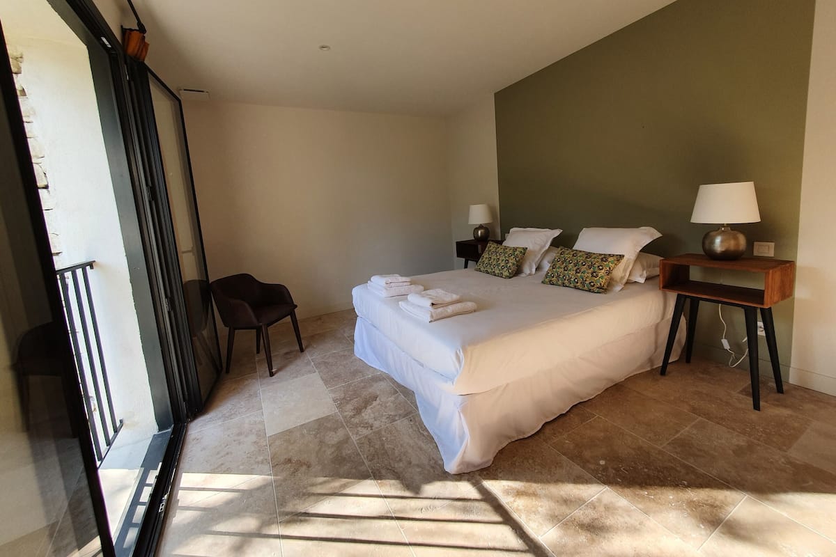 Un dormitorio con cama, dos mesitas de noche, una silla y vistas al exterior.