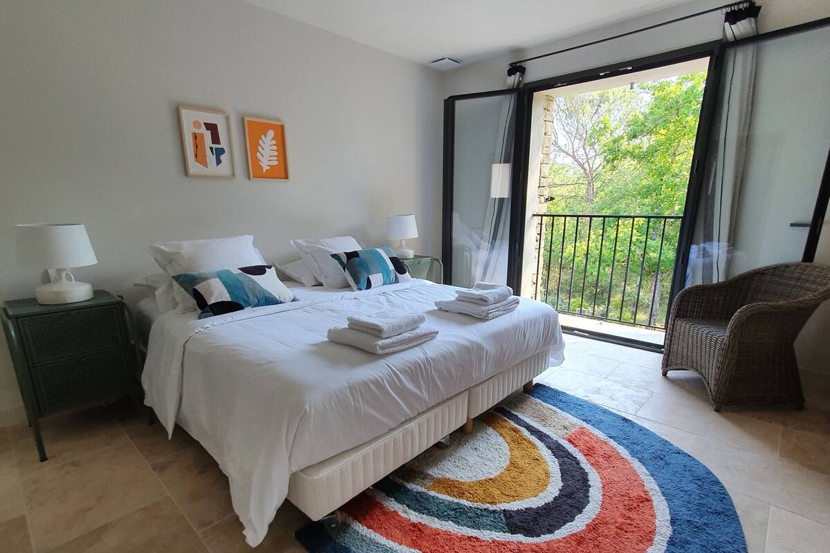 Habitación de hotel con cama, dos mesitas de noche con lámparas, alfombra colorida, una silla y balcón con vistas a zonas verdes.