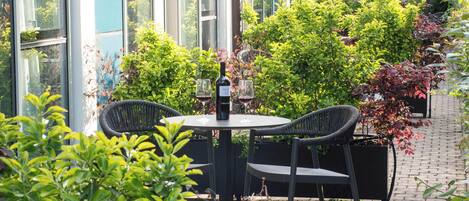 Een terras met een ronde tafel en twee stoelen, omringd door weelderige beplanting en een gebouw met grote ramen.