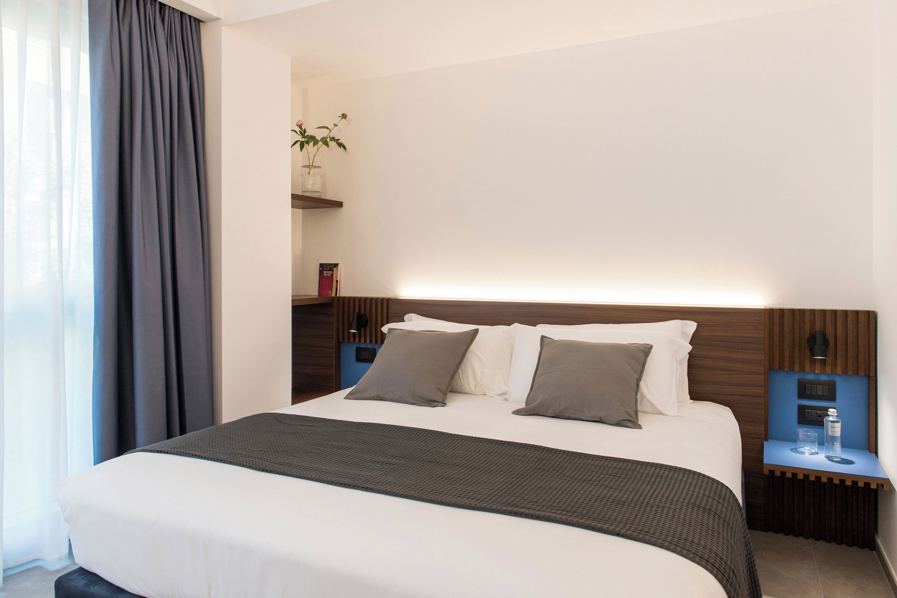 A modern hotel room with a large bed, a bedside table with a lamp, and a small shelf with a plant.