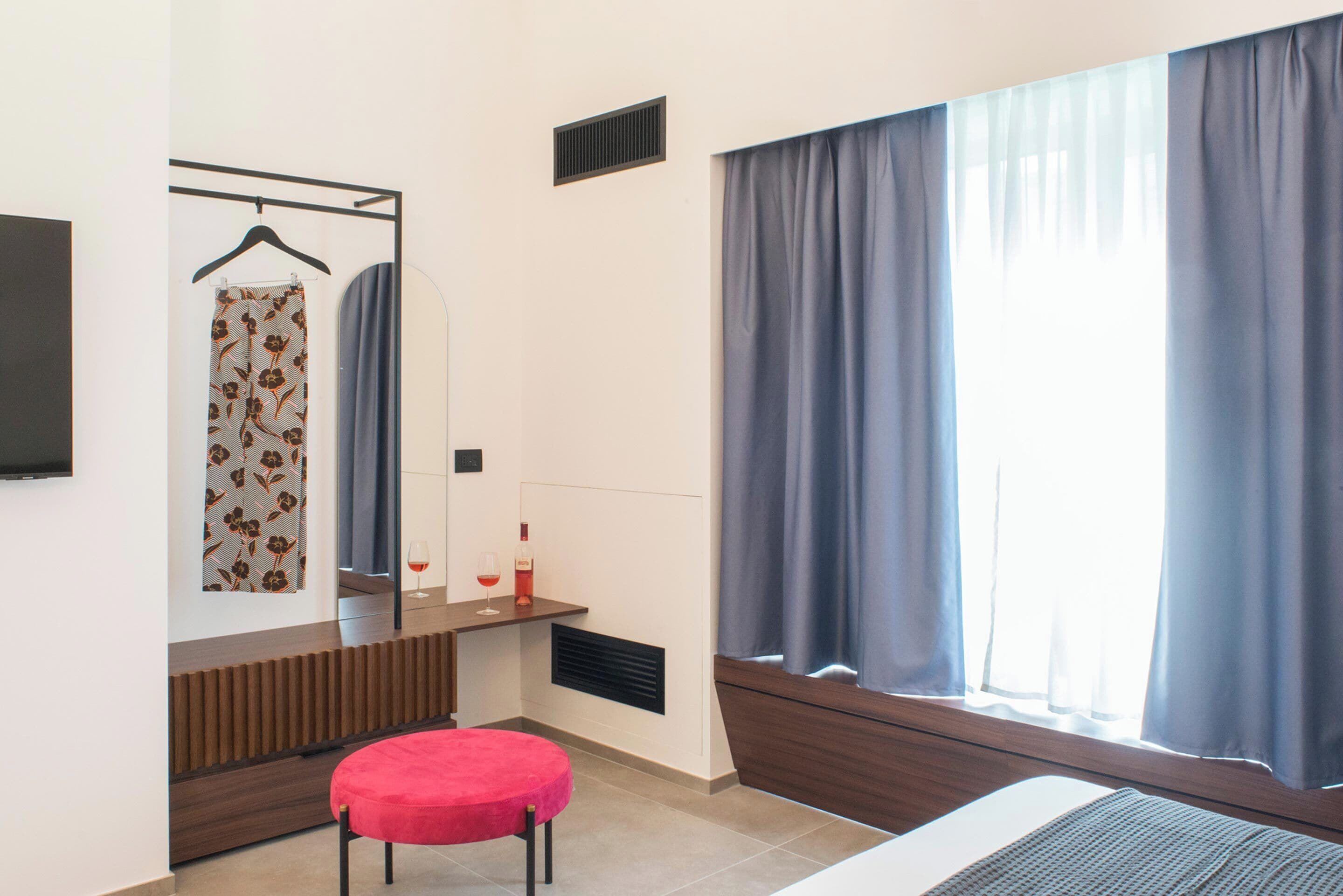 A bedroom with a bed, a red chair, a mirror, a hanging garment, and a window with curtains.