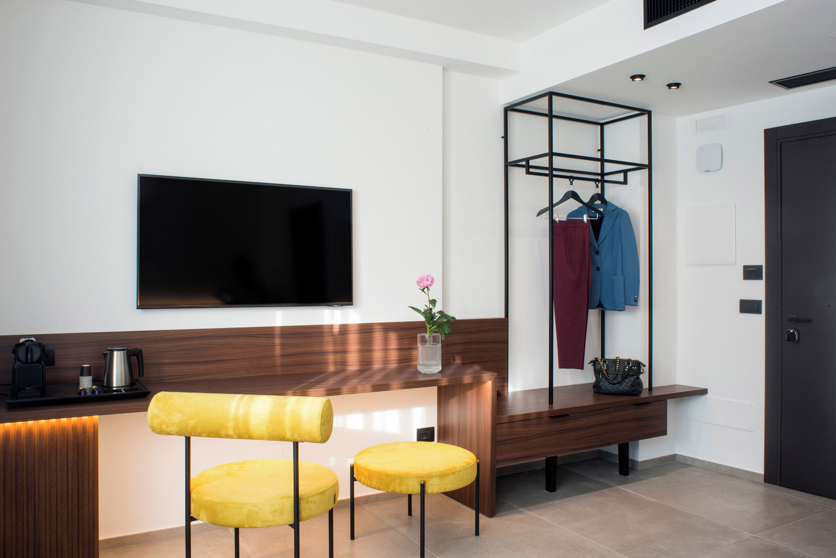 Una moderna zona de recepción de hotel con un televisor de pantalla plana, un mostrador de recepción de madera, dos sillas amarillas, un perchero con ropa y una máquina de café.
