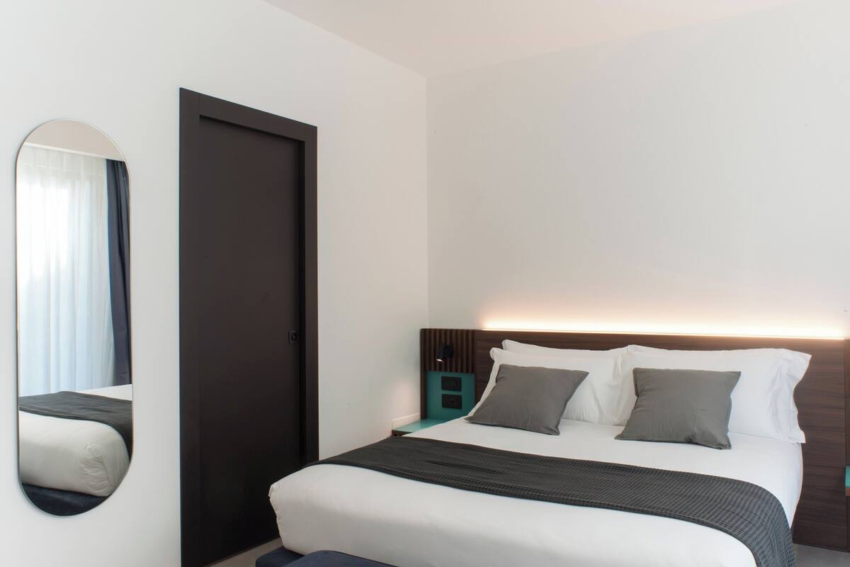 Una habitación de hotel moderna con una cama grande, cabecero oscuro, mesita de noche y un espejo.