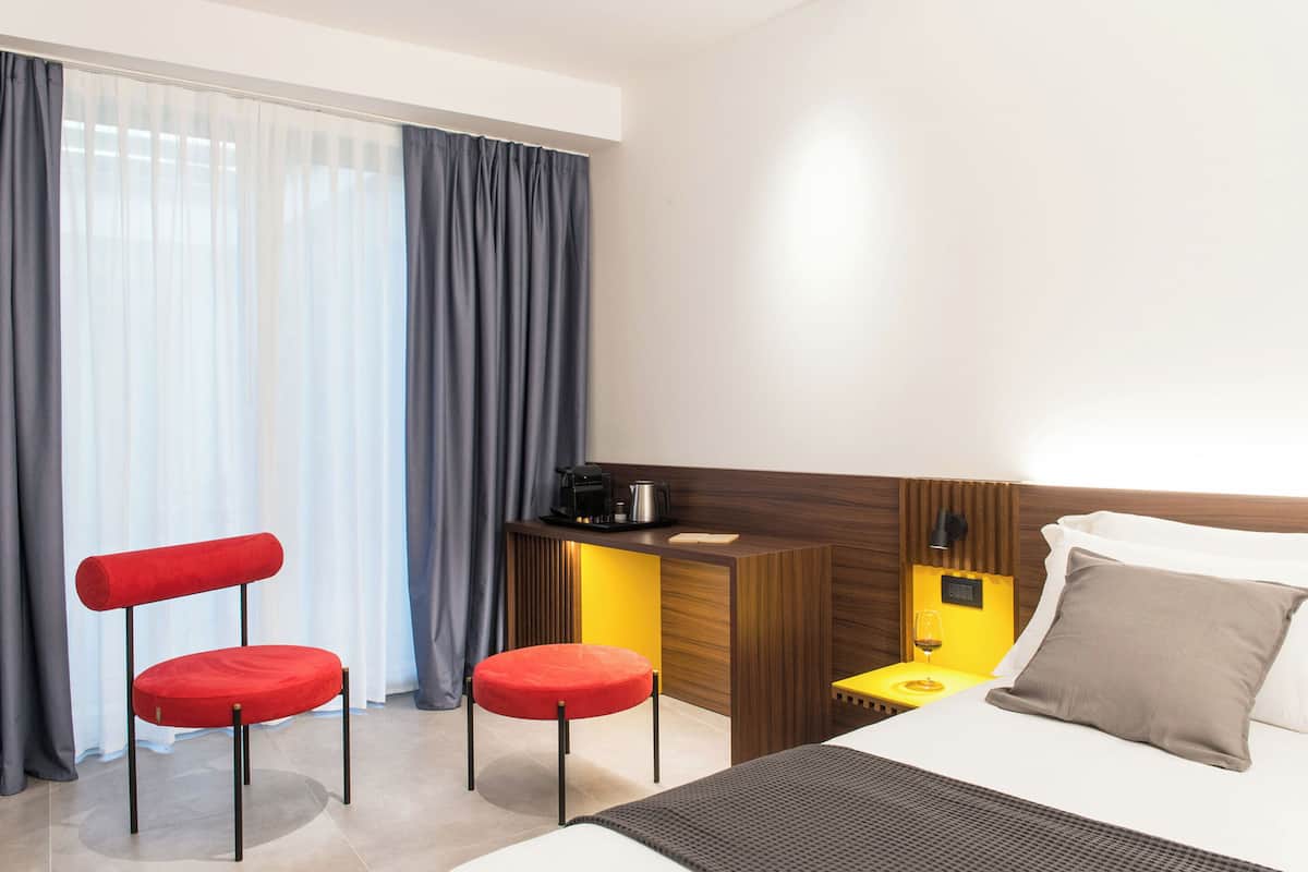 Habitación de hotel con cama, escritorio con cafetera, dos sillas rojas y mesita de noche con una copa de vino.
