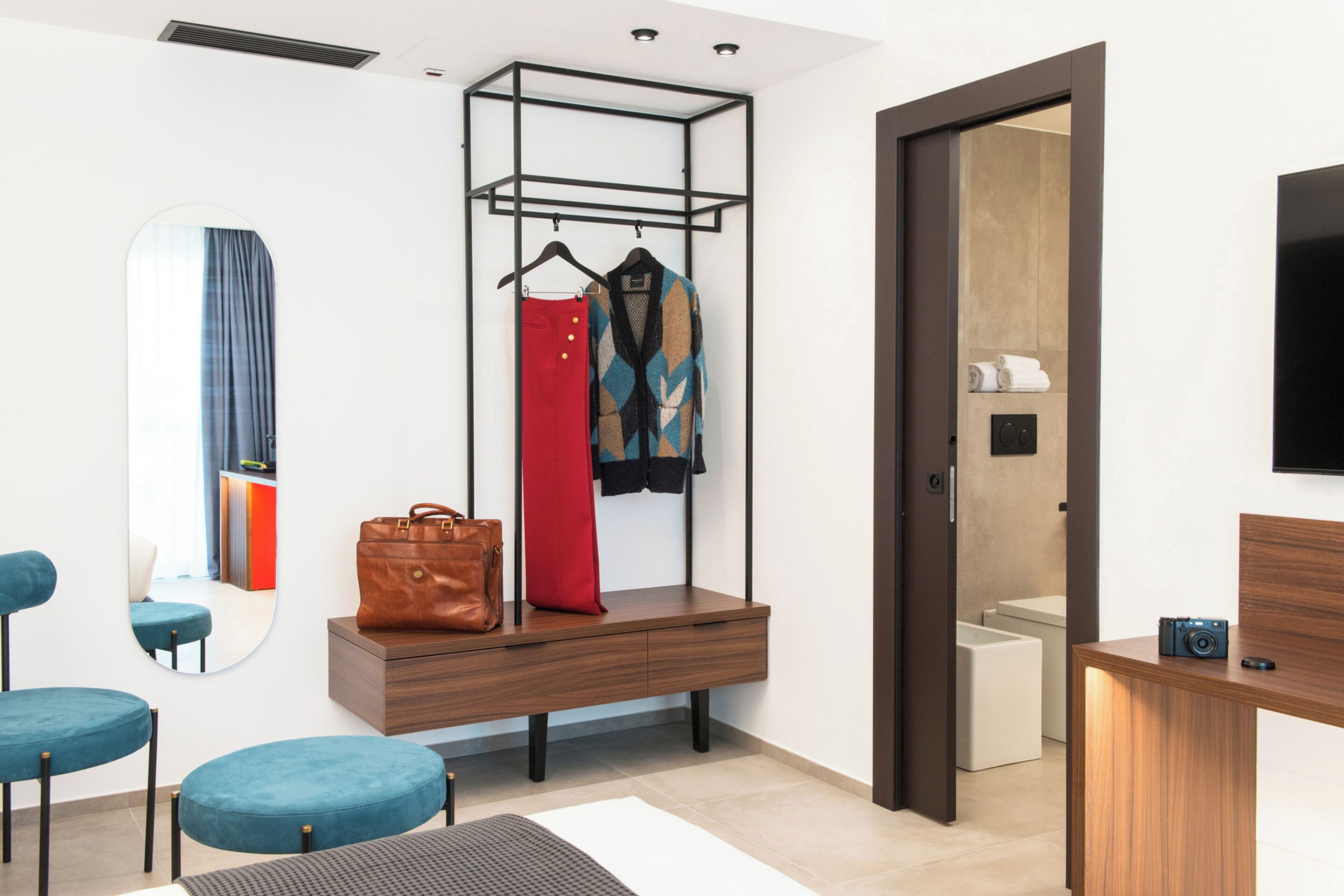 Ein modernes Hotelzimmer mit Schreibtisch aus Holz, einem Spiegel, einer Sitzecke mit blauen Stühlen, einem Kleiderständer mit Kleidung, einer braunen Ledertasche und einem Holzschrank.