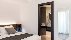 Ein modernes Hotelzimmer mit einem ordentlich bezogenen Bett, einem Nachttisch mit Lampe und einem durchgegangenen Türspalte sichtbaren Badezimmer.
