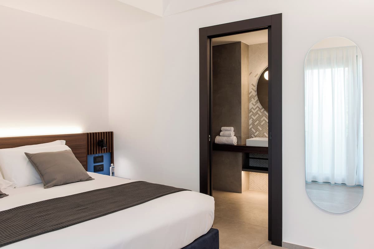 Una habitación de hotel moderna con una cama bien hecha, una mesita de noche con una lámpara y un baño visible a través de una puerta abierta.