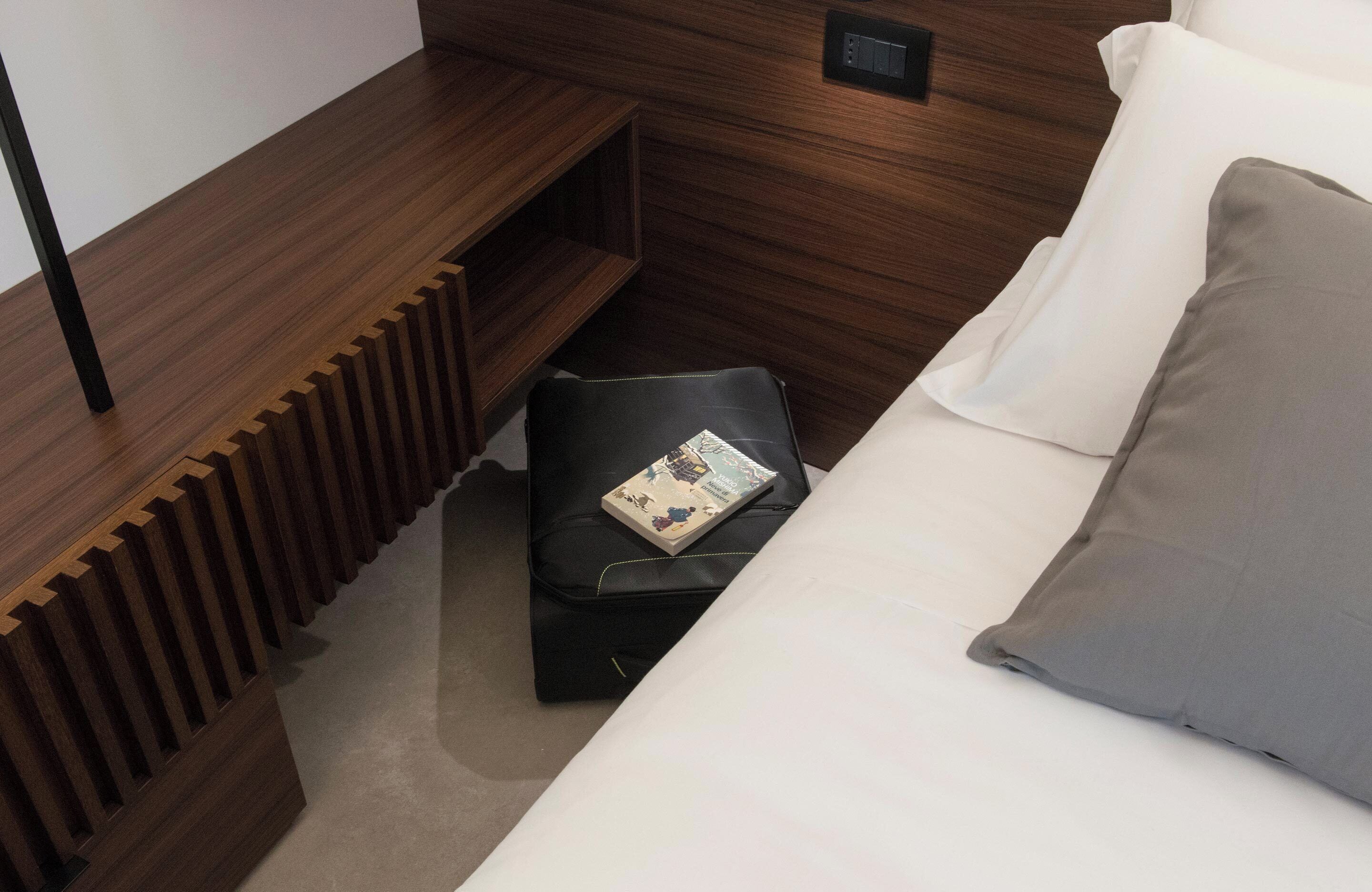 Une chambre d’hôtel bien aménagée, avec une tête de lit en bois, un lit recouvert d’une literie blanche et grise, et un petit pouf en cuir noir sur lequel repose un livre.