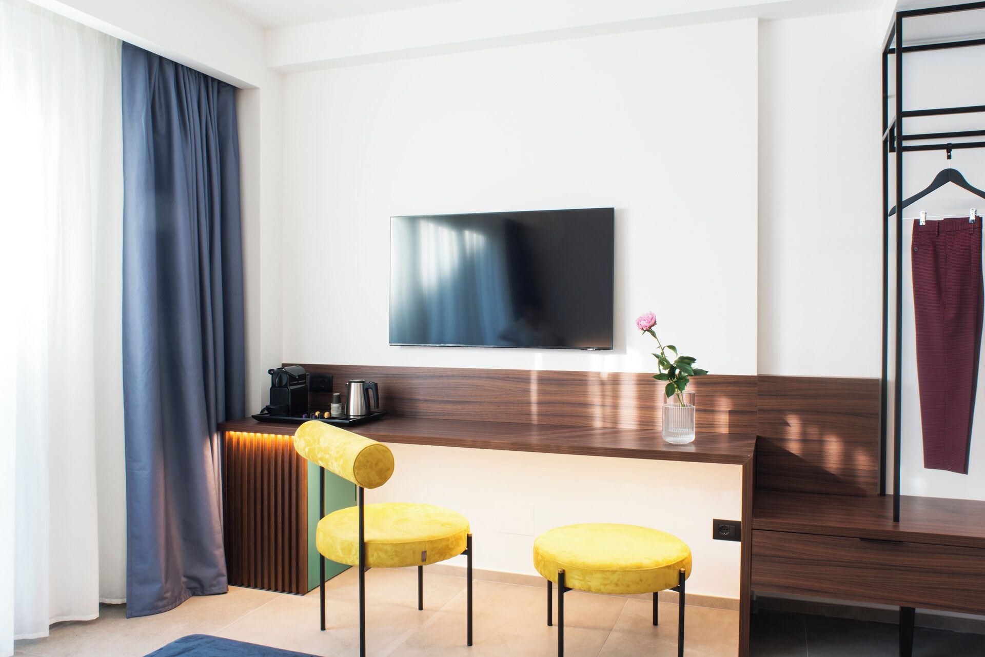 Quarto de hotel moderno com escrivaninha de madeira, duas cadeiras amarelas estofadas, televisão de tela plana na parede, cafeteira e uma pequena planta em vaso.