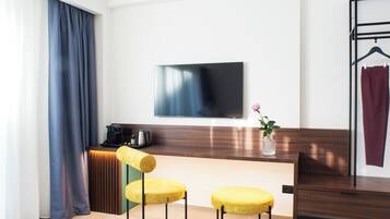 Quarto de hotel moderno com escrivaninha de madeira, duas cadeiras amarelas estofadas, televisão de tela plana na parede, cafeteira e uma pequena planta em vaso.