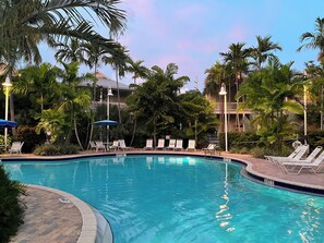 A swimming pool with lounge chairs and umbrellas, surrounded by palm trees and a building in the background. - Gone Coastal (Key West)