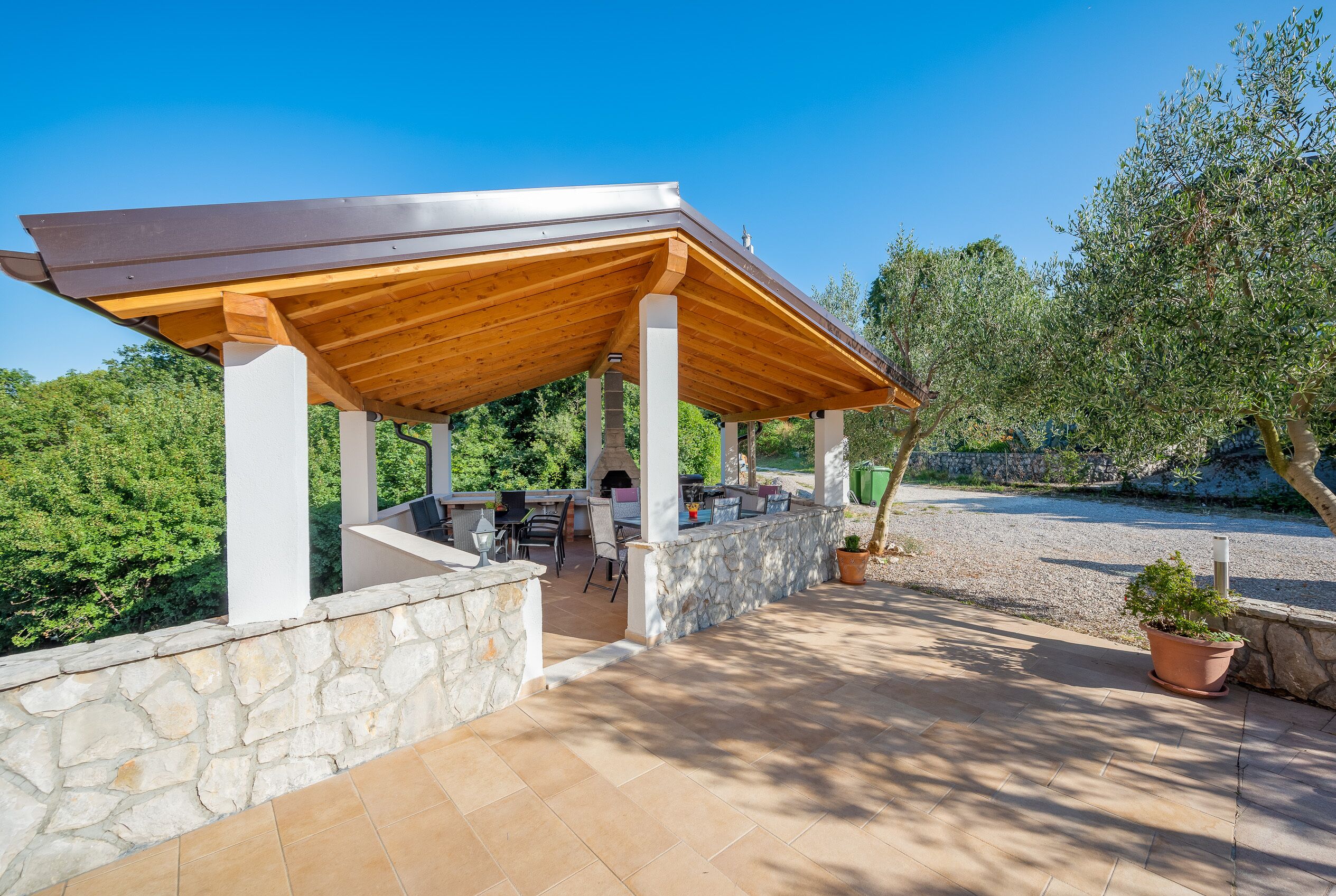 A covered outdoor area with stone walls and a wooden roof, featuring a barbecue setup and seating.