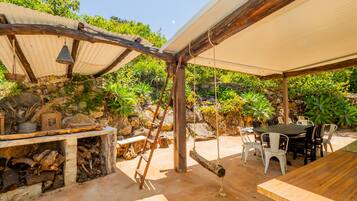 Een terras buiten met een houten tafel en stoelen, een stenen muur en diverse planten.
