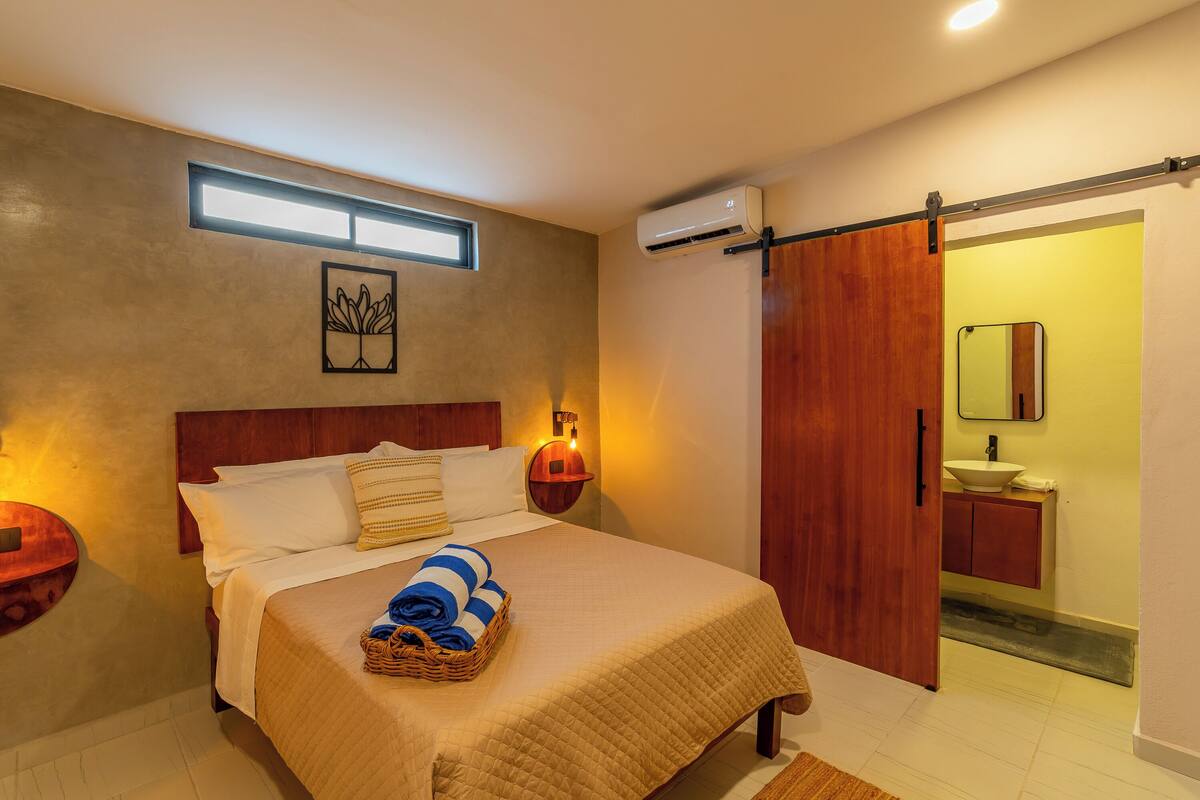 Un dormitorio con cama, lámparas de noche, aire acondicionado instalado en la pared, una puerta de madera y un baño con lavamanos y espejo.