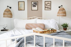 Ein ordentlich bezogenes Bett mit weißer Bettwäsche, ein gewebter Korb daneben und zwei Wandlampen.