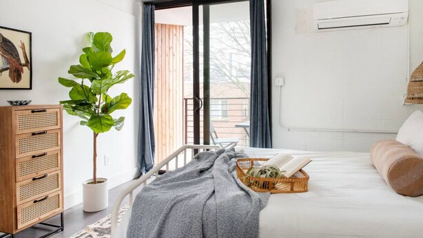 A bedroom with a bed, a bedside table, a potted plant, a window with curtains, and an air conditioning unit. - *NEW* The Jolene - Walkable | Ponce City (Atlanta)