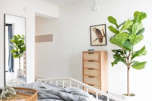 Ein Schlafzimmer mit einem Bett, einer Holzkommode, einer großen Pflanze und einem gerahmten Bild an der Wand.