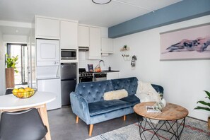 Eine moderne Küche mit weißen Schränken, einem blau-samtschwarzen Sofa, einem runden Holztisch und einem Essbereich mit einem weißen Tisch und einer Schüssel Obst.