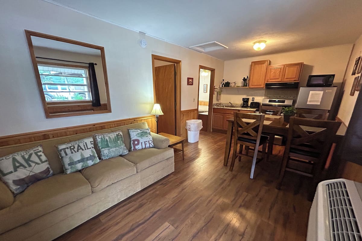 A cozy living room with a wooden floor, a beige sofa with decorative pillows, a dining table with chairs, and a kitchen area with wooden cabinets and appliances.