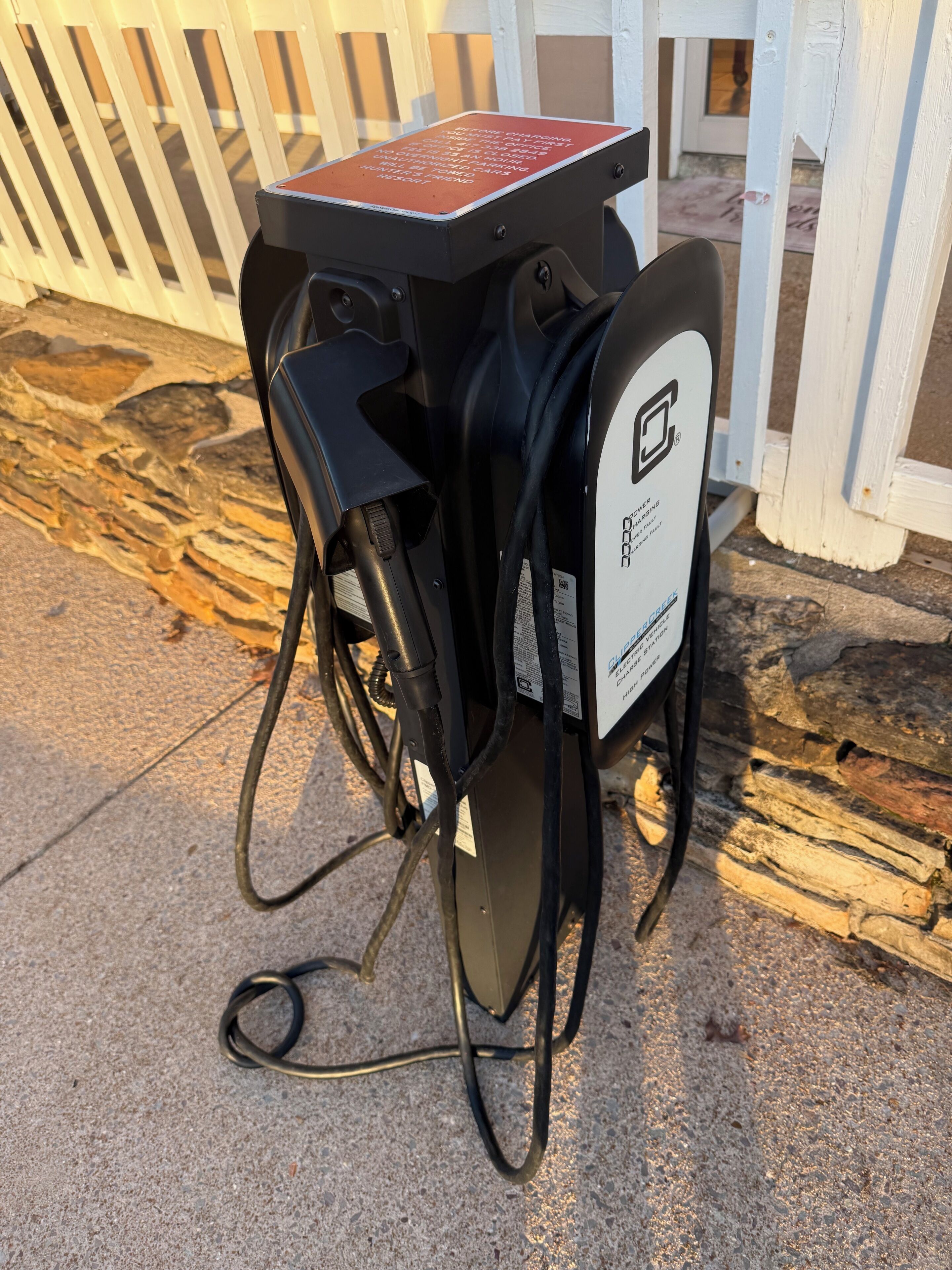 A portable gas pump with a warning sign, connected to a bicycle, on a concrete surface near a white fence.
