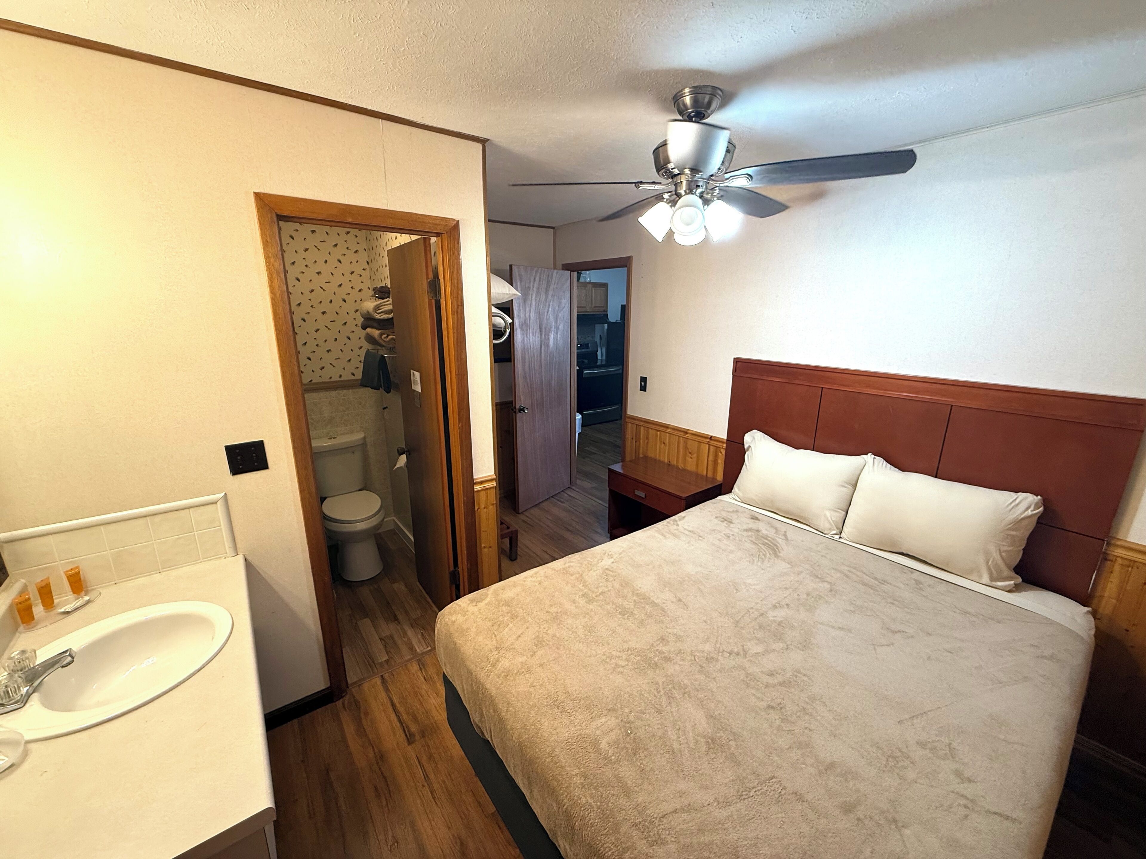 Un dormitorio con cama, ventilador de techo, cabecero de madera, baño con inodoro y lavamanos, y un pasillo que conduce a otra habitación.