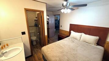 Un dormitorio con cama, ventilador de techo, cabecero de madera, baño con inodoro y lavamanos, y un pasillo que conduce a otra habitación.