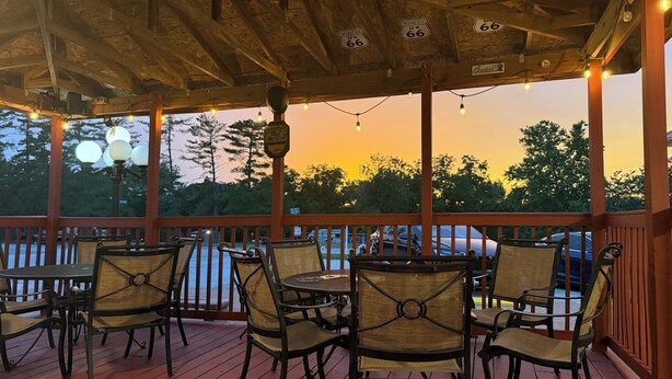 A covered outdoor seating area with wooden chairs and tables, a wooden ceiling with exposed beams, and a view of trees and a sunset.
