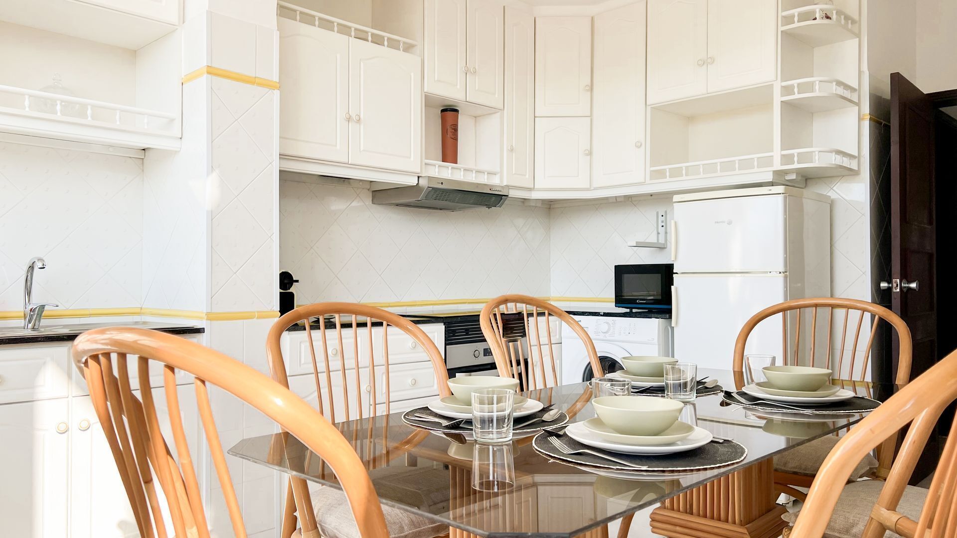 A dining area with a glass table, wooden chairs, white cabinets, and a kitchen in the background.