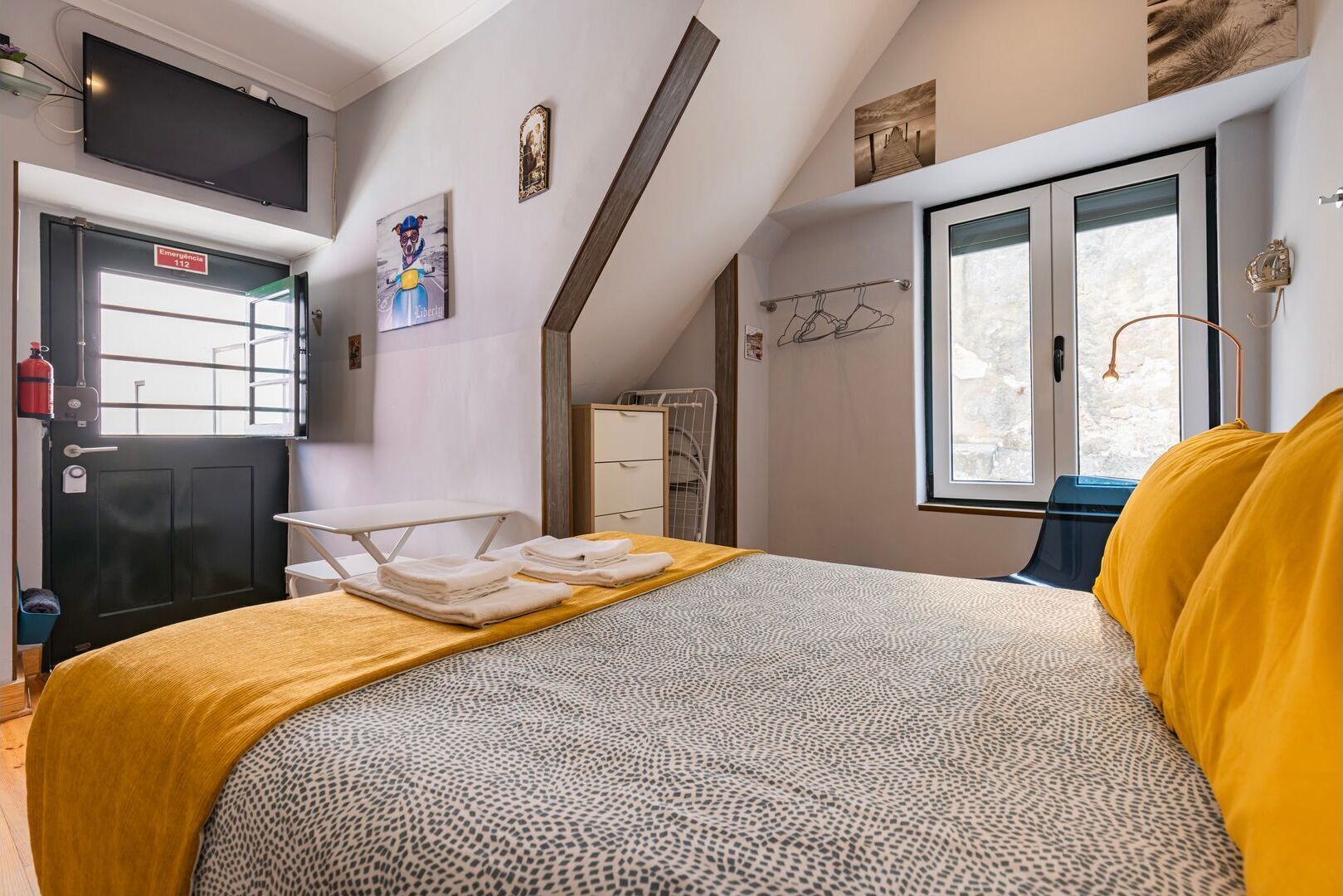 A bedroom with a bed, a window, a coat rack, and a door with an emergency exit sign.