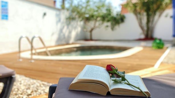 An open book with a rose on a wooden surface, a pool, and a white wall with a blue sign in the background.