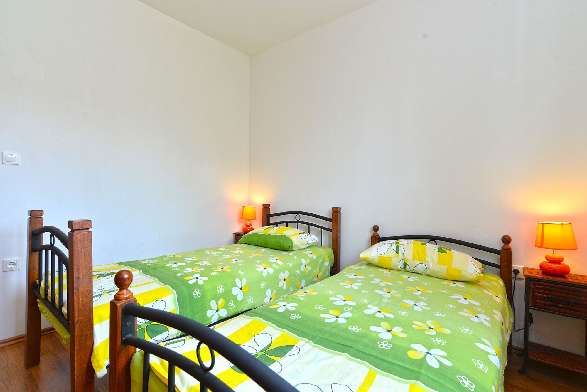 Una habitación pequeña y limpia con dos camas individuales, cada una con cabecero de madera y una mesita de noche con lámpara.